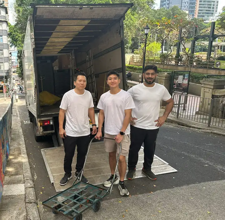 Three Brothers Moving team members in white T-shirts standing by an open moving truck with a trolley on a Hong Kong street